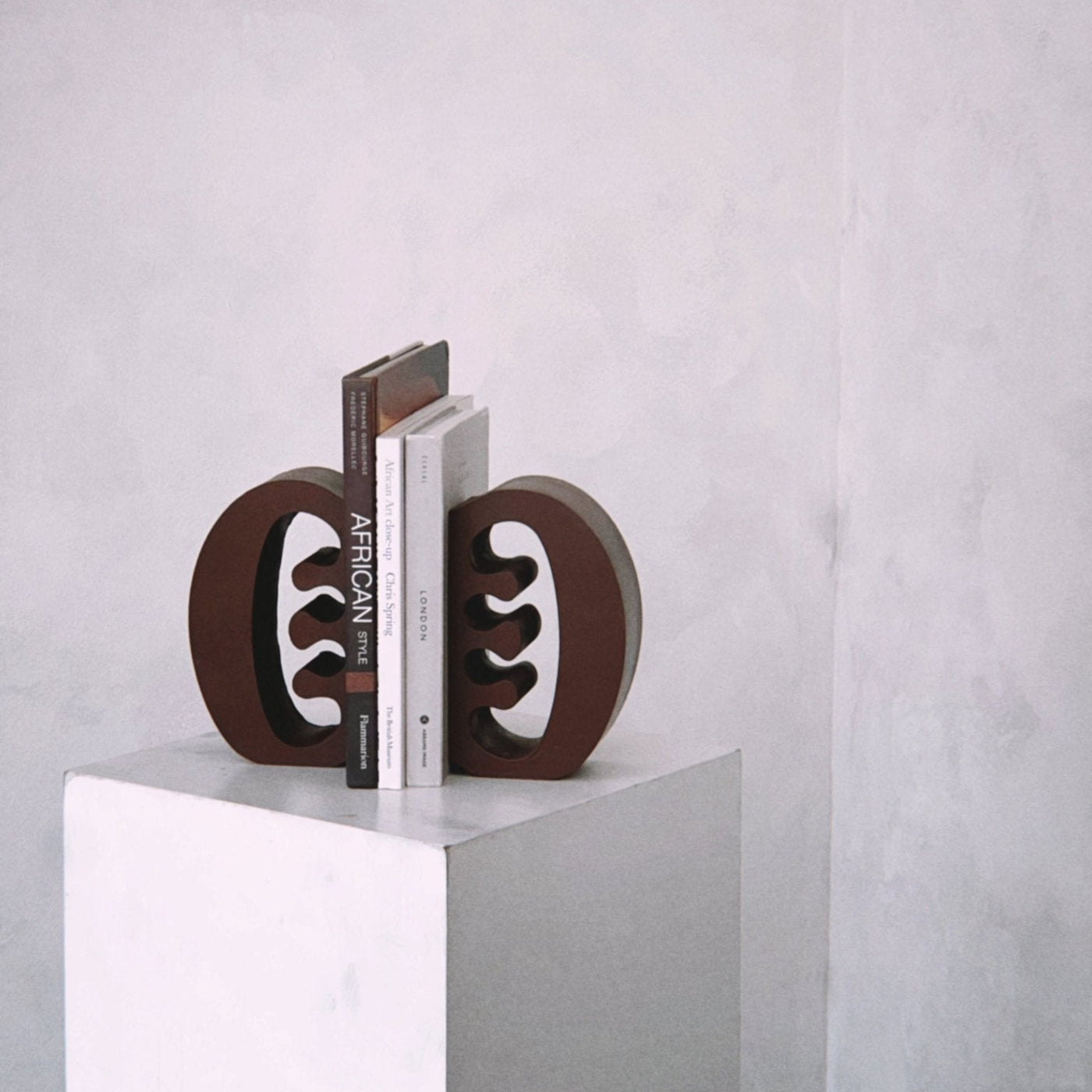 Decorative coffee coloured Ese Ne Tekrema adinkra bookends on a white plinth with a light gray lime wash background.