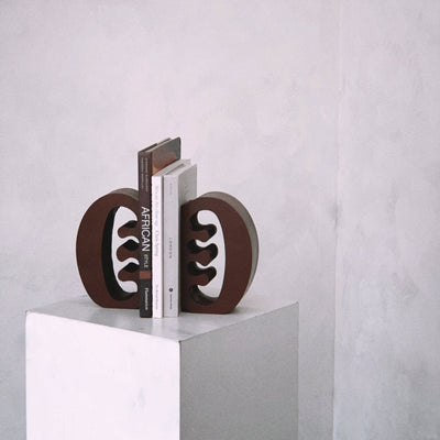Decorative coffee coloured Ese Ne Tekrema adinkra bookends on a white plinth with a light gray lime wash background.